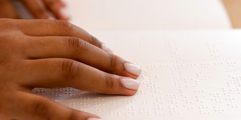 Pöydällä on kirja, jossa on pistekirjoitusta. Kirjan päällä on kirjoitusta lukevan ihmisen kädet.